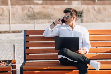 Beyaz tişörtlü, telefon ve defterli genç şık adam açık havada güneşli bir günde bankta çalışıyor. Serbest çalışıyor.