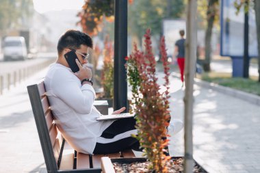 Beyaz tişörtlü, telefon ve defterli genç şık adam açık havada güneşli bir günde bankta çalışıyor. Serbest çalışıyor.