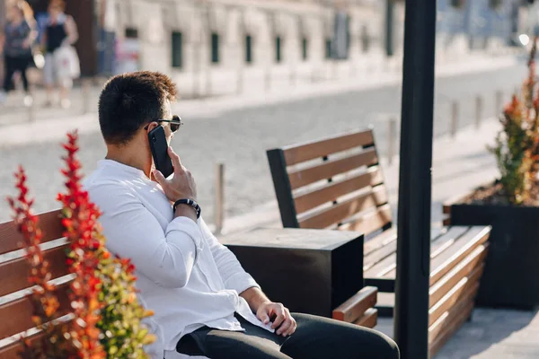 young stylish guy in white shirt with phone on bench on sunny warm day outdoors