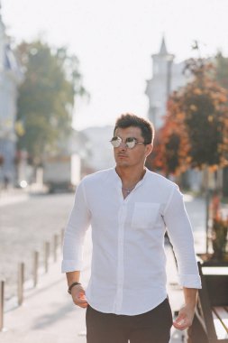 Sıcak ve güneşli bir günde Avrupa sokaklarında yürüyen beyaz tişörtlü şık bir genç.