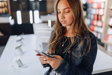 Elektronik mağazasındaki genç çekici kız masanın başında duruyor ve telefonu test ediyor. Telefon dükkanı. aygıt satın alma konsepti.
