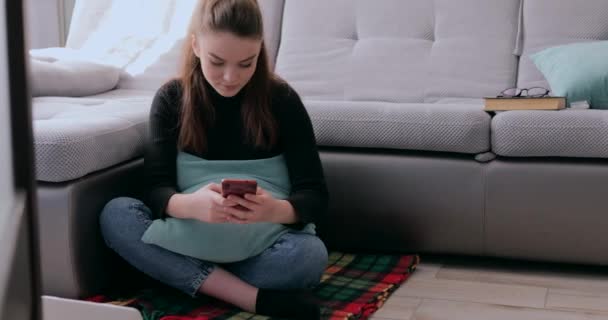 Chica bebiendo té caliente con teléfono en las manos detrás de la computadora portátil. fatiga por quedarse en casa. aislamiento y trabajo desde el hogar . — Vídeo de stock