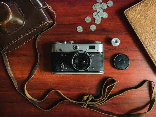 Flat lay of film camera on wooden background. Film camera with case and accessorise. Top view of film camera, case, coins, notebook. Concept of photographer or traveler equipment kit.