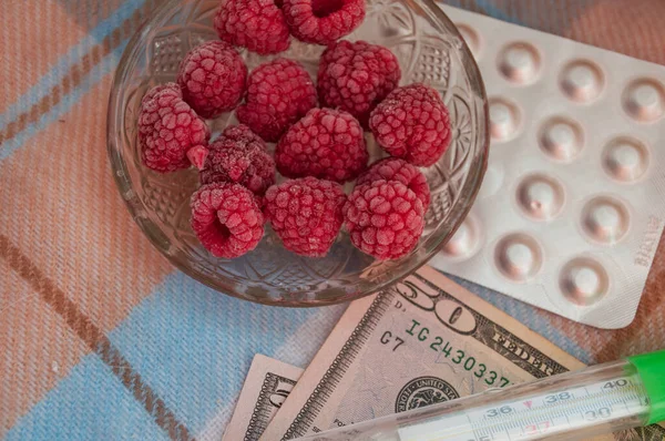 how much it costs to treat flu. frozen raspberries on a transparent saucer, thermometer, money and tablet.