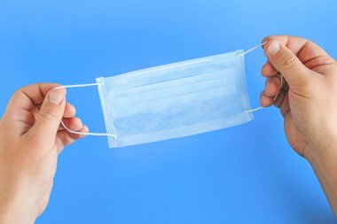 Mavi arka planda erkek eli takmış tıbbi bir maskenin yakın plan görüntüsü. Birinci şahıs görüşü. Uygun kullanım ve virüs koruması.