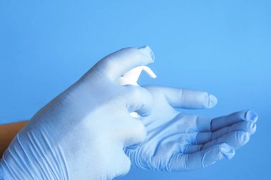 Dezenfektanı nasıl kullanacağının resmi. Mavi arka planda lateks koruyucu eldivenlerle el ele tutuşan antiseptik bir konteynır. Virüse, tıbba ve sağlığa karşı korunma kavramı.