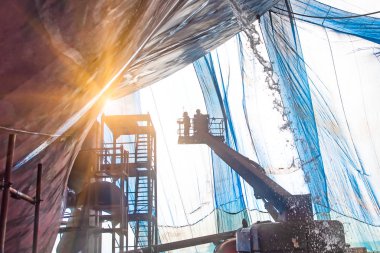 Steel basket carry by cherry picker crane for gang of worker power man, painting man or brush man working in high position, working at risk in dust under mesh sheet protectio