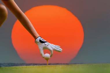 hand of woman golfer holding golfball to marking point to being putting on the green of golf course, large sunset drop to the sea in background