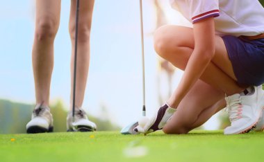 Golf topu tutan genç bayan golfçünün eli ahşap tişörte uzanmış topa vurmaya hazırlanıyor ve vurmaya hazırlanıyor. Rakip rakip rakip ya da golf arkadaşı arka planda izliyor.