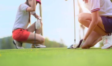 Golf topu tutan genç bayan golfçünün eli ahşap tişörte uzanmış topa vurmaya hazırlanıyor ve vurmaya hazırlanıyor. Rakip rakip rakip ya da golf arkadaşı arka planda izliyor.