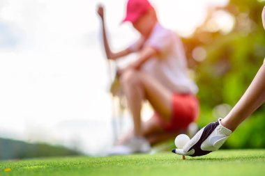 Golf topu tutan genç bayan golfçünün eli ahşap tişörte uzanmış topa vurmaya hazırlanıyor ve vurmaya hazırlanıyor. Rakip rakip rakip ya da golf arkadaşı arka planda izliyor.