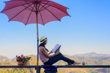 Kadın gezgin rahat bir şekilde terasta oturur, sabah kitap okur, yaz tatilinde seyahat eder, seyahatin tadını çıkarır ve hareketli bir şekilde dinlenir.,