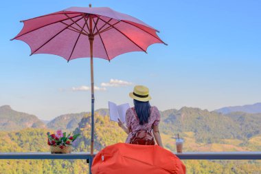 Kadın gezgin rahat bir şekilde terasta oturur, sabah kitap okur, yaz tatilinde seyahat eder, seyahatin tadını çıkarır ve hareketli bir şekilde dinlenir.,