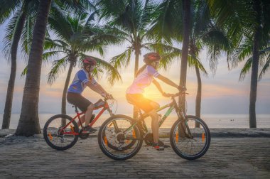 Couple lover enjoy riding bicycle under coconut palm trees at light of sunset, relax and comfortable at the end of the day together