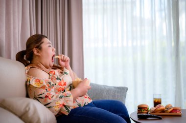 Şişko kadın evde abur cubur yerken televizyon izlemekten zevk alıyor.
