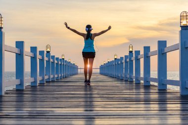 Spor kadını, gün batımında ahşap köprüde egzersiz yapıyor. Denizde iskelede çalışırken müziğin keyfini çıkarıyor.