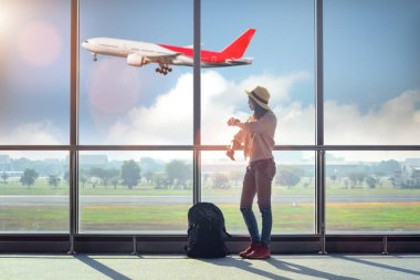 Kadın turist ya da gezgin, havaalanı terminalinin ulaşım koridorunda endişeli ve üzgün bir şekilde, yurtdışına gitmek için uçak kalkışını, arka plandaki uçuş uçağının inişini bekliyor.