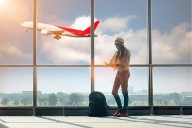 Kadın turist ya da gezgin, havaalanı terminalinin ulaşım koridorunda endişeli ve üzgün bir şekilde, yurtdışına gitmek için uçak kalkışını, arka plandaki uçuş uçağının inişini bekliyor.