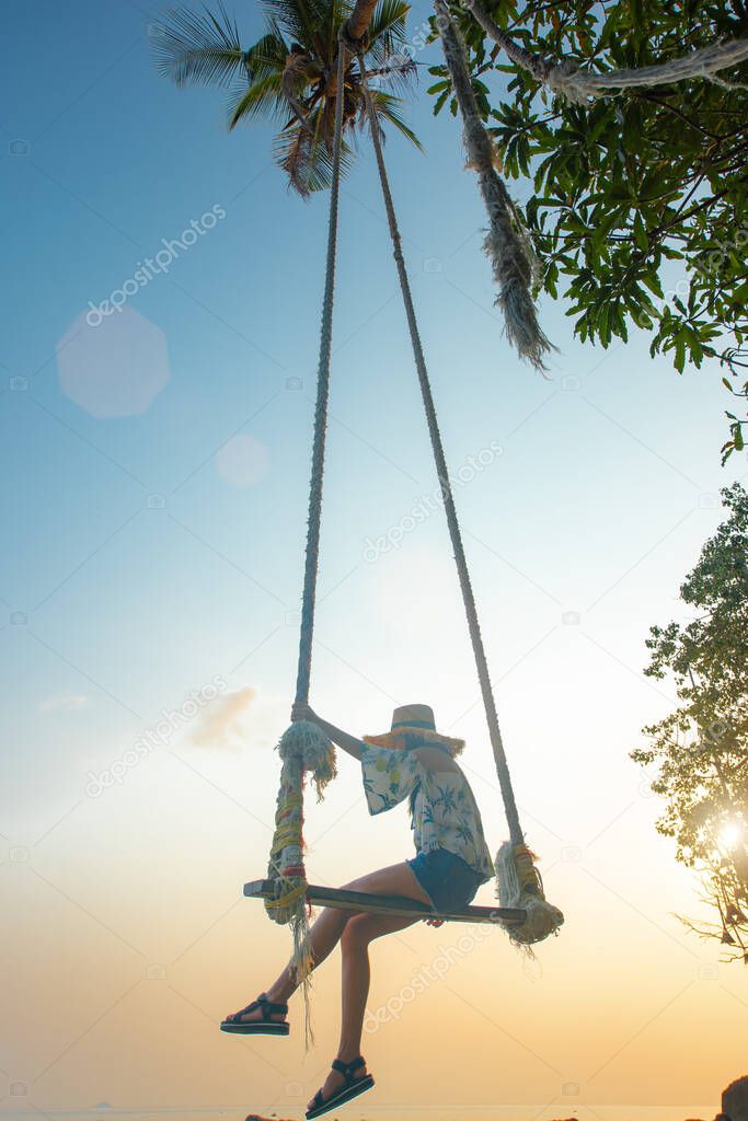 young woman sitting on sway the wooden swing under coconut palm tree and shadow of the trees over the sea beach, sunbeam and sunset light feel comfortable and most relaxation