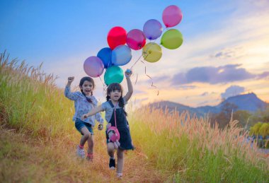 Tepelerdeki çayırlarda elinde renkli balonlarla koşan ikiz çocuklar.