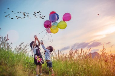 Heyecanlı ve mutlu bir şekilde balonla çayırlarda koşan çocukları oynuyorum. Gün batımı ve arka planda kuş sürüsü var.