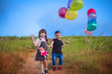 Çocuklar kız ve erkek el ele tutuşup tepeden aşağı inen balonlarla, tepe boyunca uzanan çayır tarlasıyla