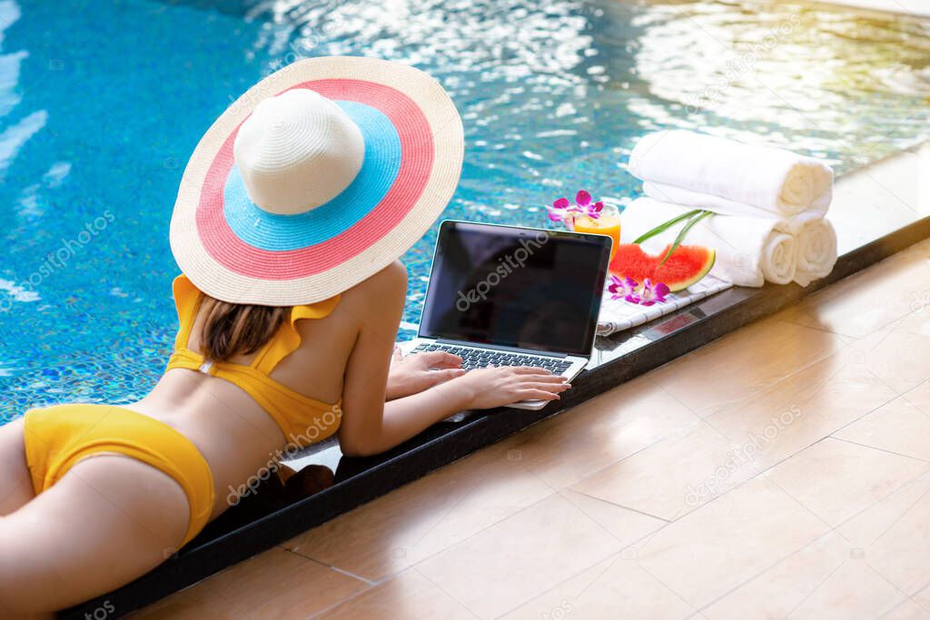 Hand of woman playing on the keyboard of notebook computor at the edge of swimming pool, enjoy with orange juice and water melon fruit while relax in summer time vacation 