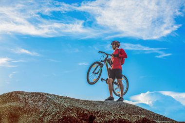 Güneşli bir günde mavi gökyüzüne karşı kayalık arazide bisikletini taşıyan bisikletçi.