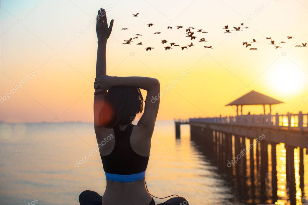 young sporty woman perform in yoga practice on wooden bridge in the sea at sunset, with flock of bird flying over the water in background