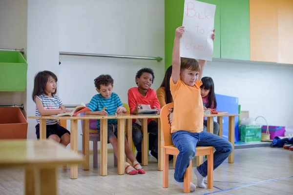 Head of the class in preschool present paper poster reading as Back to school in classroom with group of friends and teacher kindergartner stay in row background