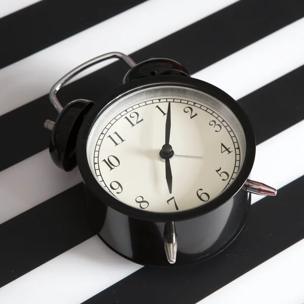 Μαύρο ρολόι συναγερμών σε ένα μαύρο και άσπρο ριγέ χαρτοπετσέτα δείχνει 7 η ώρα στο κομοδίνο — Φωτογραφία Αρχείου