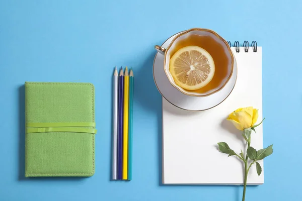 Fyrkantig anteckningar på fjädrar med vitt kraftpapper, penna, gul ros och kopp te är på en blå bakgrund. — Stockfoto