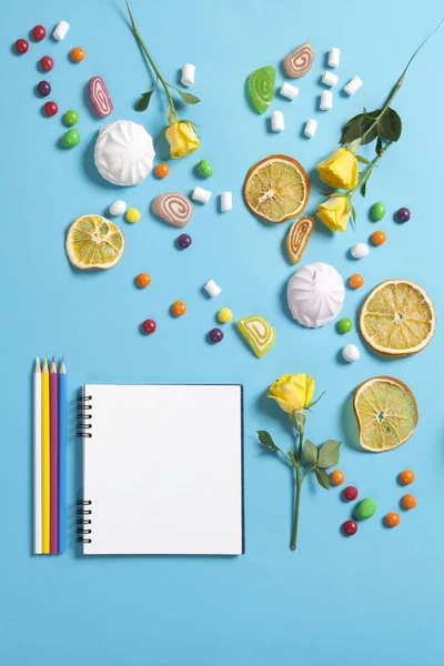 Marshmallows, godis, gelé bönor, sötsaker, torr muggar apelsiner och gula rosor faller på ett anteckningsblock på en blå bakgrund. Advertising Space — Stockfoto
