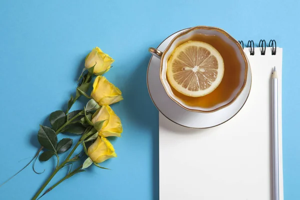 Bloc de notas cuadrado en muelles con papel kraft blanco, lápiz, rosa amarilla y taza de té están sobre un fondo azul . —  Fotos de Stock