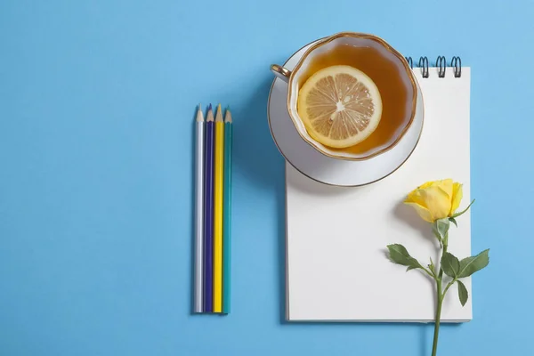 Fyrkantig anteckningar på fjädrar med vitt kraftpapper, penna, gul ros och kopp te är på en blå bakgrund. — Stockfoto