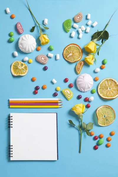 Marshmallows, doces, geléias, doces, canecas secas laranjas e rosas amarelas caindo em um bloco de notas em um fundo azul. Espaço de publicidade — Fotografia de Stock