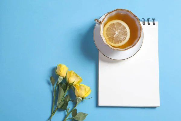 Fyrkantig anteckningar på fjädrar med vitt kraftpapper, gul ros och kopp te är på en blå bakgrund. — Stockfoto