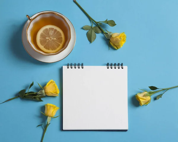 Mavi bir arka plan üzerinde beyaz kraft kağıt, Sarı Gül ve çay ile yaylar üzerinde kare not defteri var. Boşaltmak — Stok fotoğraf