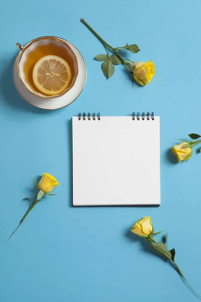Mavi bir arka plan üzerinde beyaz kraft kağıt, Sarı Gül ve çay ile yaylar üzerinde kare not defteri var. Boşaltmak — Stok fotoğraf