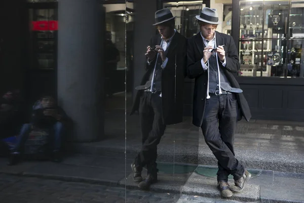 Street actor magician in a hat and a strict suit is leaning against a mirror display. — Stock Photo, Image