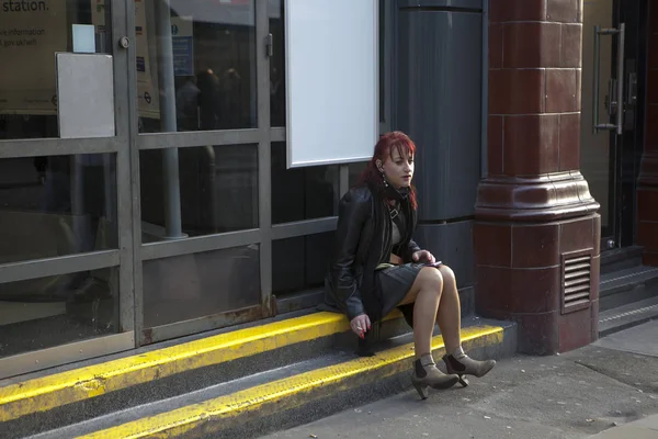 A lonely woman wears a short green skirt and a brown leather jacket, sits on the curb and smokes a cigarette — Stock Photo, Image