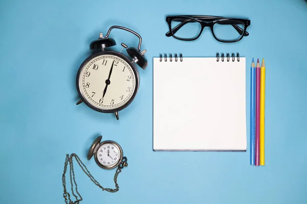 Klocka, vit anteckningsbok på fjädrar, Glasögon och färgpennor på en blå bakgrund — Stockfoto