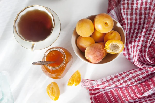 Les abricots dans un bol et la confiture dans un bocal sur une table blanche avec une serviette rouge et blanche, des blancs d'été faits à la main — Photo