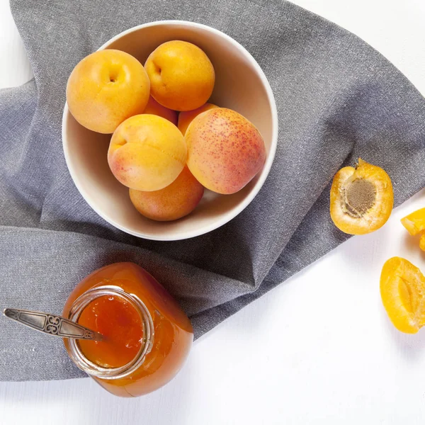 Les abricots dans un bol et la confiture dans un bocal sur une table blanche avec une serviette rouge et blanche, des blancs d'été faits à la main — Photo