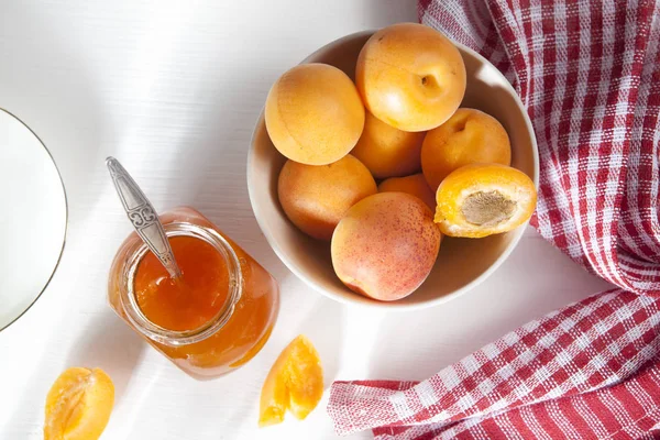 Les abricots dans un bol et la confiture dans un bocal sur une table blanche avec une serviette rouge et blanche, des blancs d'été faits à la main — Photo