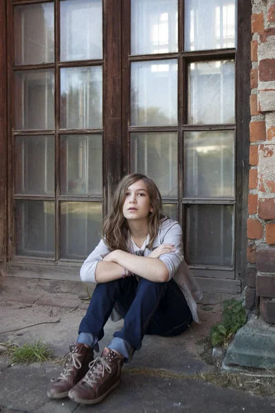 Sad girl with long hair in jeans of thirteen sits near big window — Stock Photo, Image