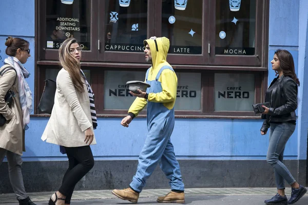 Passers-by andando ao longo da rua. Um mendigo num fato amarelo e um chapéu na mão pedem dinheiro. — Fotografia de Stock