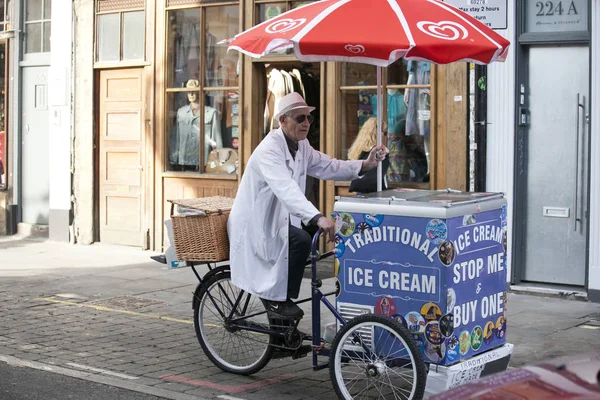 Un vendedor de helados de edad avanzada en una bata blanca monta en bicicleta con su carrito de helados —  Fotos de Stock