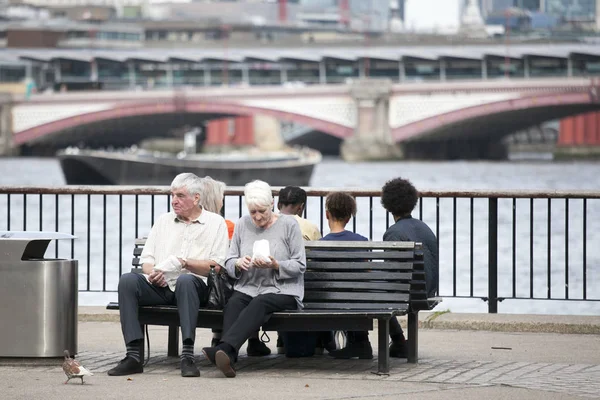 An elderly couple eating popcorn, sitting on a bench overlooking the Thames. South Shore of the Thames — Stock Photo, Image