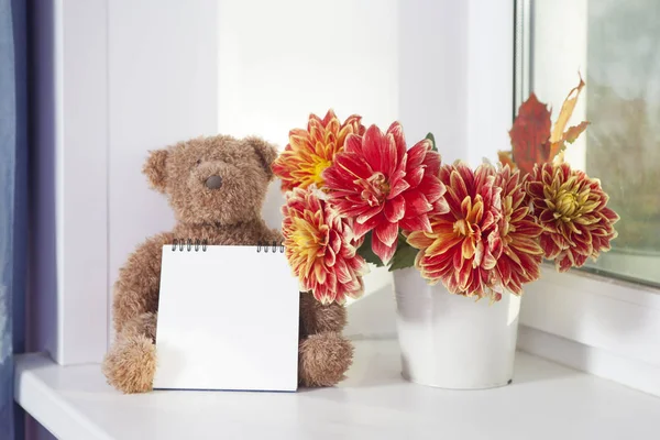 The teddy-bear next to bouquet of autumn dahlias. The teddy-bear holds a notepad for writing. — Stock Photo, Image